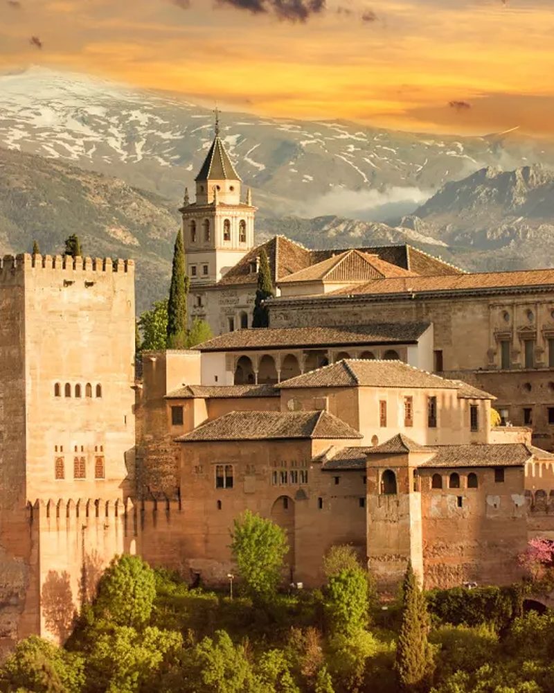 Castelo histórico na Espanha com montanhas ao fundo, iluminado pela luz dourada do entardecer