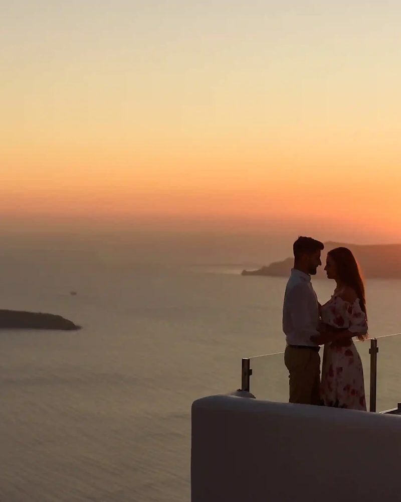 Casal abraçado admirando o pôr do sol em terraço com vista para o mar em Santorini